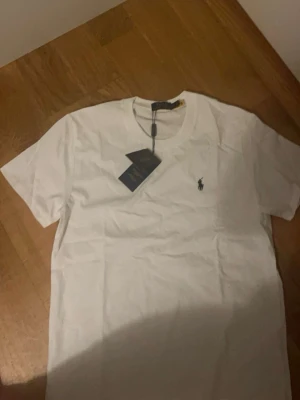 Vit t-shirt från Polo Ralph Lauren - Klassisk vit t-shirt från Polo Ralph Lauren med den ikoniska broderade loggan på bröstet. T-shirten har rund hals och korta ärmar, perfekt basic-plagg till garderoben. Tillverkad i mjuk bomull för skön känsla. Kontakta vid minsta fråga!
