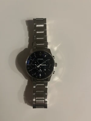 Citizen kronografklocka i silver - Stilren kronografklocka från Citizen med svart urtavla och flera små urtavlor. Armbandet är i borstat och polerat silverfärgat stål och har en klassisk länkdesign. Klockan har tydliga indexmarkeringar och två tryckknappar vid sidan av boetten.