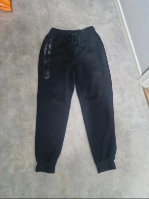 Svarta Nike mjukisbyxor med dragsko - Svarta mjukisbyxor från Nike med dragsko i midjan och resår vid bensluten. Byxorna har en snygg detalj med svart tejp längs ena fickan och diskret Nike-logga på låret. Perfekta för en avslappnad och sportig stil.