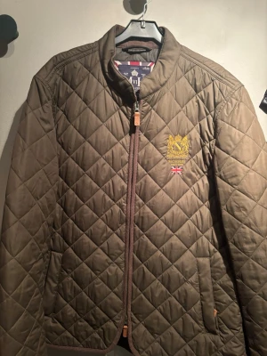 Quiltad brun jacka från Morris - Snygg quiltad brun jacka från Morris med gul brodyr och brittisk flagga på bröstet. Jackan har dragkedja framtill, ribbade detaljer och två fickor. Perfekt för dig som gillar klassisk stil med modern twist.