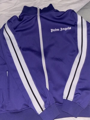 Lila Palm Angels hoodie med vita ränder - Säljer en lila hoodie från Palm Angels med vita och svarta ränder längs ärmarna. Dragkedja framtill och hög krage. Ikonisk Palm Angels-logga på bröstet. Perfekt för dig som gillar streetwear och vill sticka ut med en snygg färg.