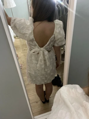 Studentklänning - Supersöt vit kort klänning med puffärmar och öppen rygg. Klänningen har ett diskret blommigt mönster, knyt i ryggen och volangdetaljer. Perfekt för studenten.