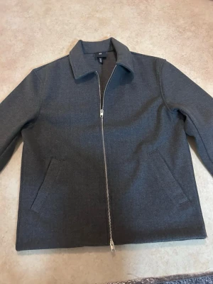 Grå jacka med dragkedja från H&M - Snygg grå jacka från H&M med klassisk krage och hel dragkedja framtill. Jackan har två snedställda fickor och är i ett mjukt material som passar perfekt till vardagslooken. Enkel och stilren design som är lätt att matcha.