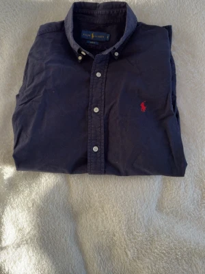 Mörkblå skjorta från Ralph Lauren - Klassisk mörkblå skjorta från Ralph Lauren med knappar framtill och röd broderad logga på bröstet. Skjortan har lång ärm, button down-krage och är tillverkad i mjuk bomull. Perfekt för en stilren och avslappnad look.