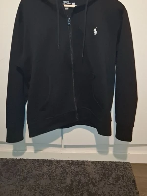 Svart hoodie från Polo Ralph Lauren - Klassisk svart hoodie från Polo Ralph Lauren med dragkedja framtill och vit broderad logga på bröstet. Hoodien har huva med dragsko och två fickor fram. Tillverkad i mjukt bomullsmaterial som känns skönt mot huden.