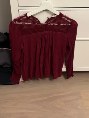 Vinröd blus med spetsdetaljer - Snygg vinröd blus med långärmad design och transparenta spetsdetaljer upptill. Blusen har rund halsringning och ett lätt plisserat tyg som ger ett flowy intryck. Perfekt för dig som vill ha något extra till outfiten.