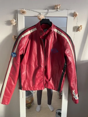 Röd skinnjacka med vita ränder - Snygg röd skinnjacka med vita och svarta ränder längs ärmarna. Jackan har ståkrage med tryckknapp, dragkedja framtill och coola patchar på bröstet och ena ärmen.två dragkedjor på varje arm och två sidor där midjan är har dragskedjor . Perfekt för dig som vill sticka ut med en sportig och retroinspirerad look.