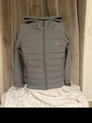 Lacoste jacka - Snygg grå pufferjacka från Lacoste med huva och dragkedja framtill. Jackan har quiltade partier och den klassiska Lacoste-loggan på bröstet. Perfekt för kyligare dagar och har en modern, clean look. Har en liten liten fläck på framsidan…