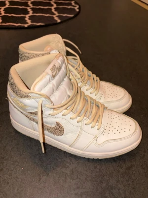 Nike Air Jordan 1 Mid vita sneakers - Snygga Nike Air Jordan 1 Mid sneakers i vitt skinn med beige detaljer och snörning. Swoosh-loggan och hälpartiet har ett unikt glittrigt mönster som sticker ut. Perfekta för dig som vill ha en stilren men ändå unik look.