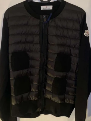 Svart cardiganjacka från Moncler - Snygg svart cardiganjacka från Moncler med quiltad framsida och stickade ärmar. Jackan har rund halsringning, dragkedja framtill och två stora fickor. Moncler-logga på ärmen. Perfekt för lager på lager och streetstyle. Orderbekrätelse följs med! Helt ny, säljs för den inte passade så bra! De flesta i marknaden säljer lågt på grund av att de inte är autentiska!
