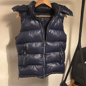 Mörkblå dunväst med huva från Leon Marcél - Snygg mörkblå dunväst från Leon Marcél med avtagbar huva och två dragkedjeförsedda fickor. Västen har en glansig finish och är riktigt snygg för både en utstickande och stilren look. Perfekt till våren och kyliga sommar kvällar! Västen är sprillans ny och har prislapp mm kvar!