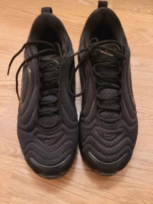 Nike Air Max 720 svarta sneakers - Nike Air Max 720 sneakers i helsvart med vågigt mönster och synlig Air-sula. Skorna har en sportig look med mesh- och syntetmaterial, samt diskreta Nike-loggor på sidan och tån. Låga med snörning och dragflik bak. Perfekta för en streetwear-stil.