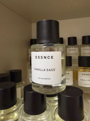 ESSNCE Vanilla Sass' Eau de Parfum - En stilren parfymflaska i glas med svart lock från ESSNCE. Doften heter Vanilla Sass' och är en Eau de Parfum. Flaskan är genomskinlig med en vit etikett och minimalistisk design. Perfekt för dig som gillar vaniljtoner och modern estetik.