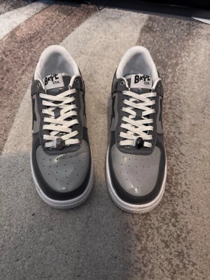 Grå Bape Sta sneakers - Säljer ett par grå Bape Sta sneakers med svart stjärndetalj på sidan. Skorna har vit sula, vita skosnören och är tillverkade i glansigt lädermaterial. Använda 3 gånger  