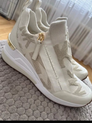 Vita Michael Kors sneakers med gulddetaljer - Snygga vita sneakers från Michael Kors med gulddetaljer på hälen och dragkedja på sidan. Skorna har stickat tyg med diskret logomönster och inslag av beige. Bekväm platåsula och modern design som sticker ut.