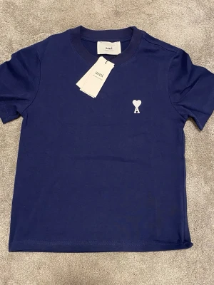 Ami Paris tröja - Snygg blå t-shirt från Ami med klassisk passform och korta ärmar. T-shirten har en vit broderad hjärtdetalj på bröstet och är gjord i mjuk bomull. Perfekt för en stilren och avslappnad look. Storlek: 164cm.