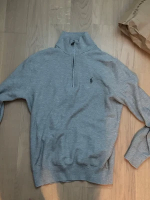 Grå halvzip från Polo Ralph Lauren - Snygg grå stickad tröja från Polo Ralph Lauren med halv dragkedja och hög krage. Tröjan har lång ärm, ribbade muddar och den klassiska broderade loggan på bröstet. Perfekt för lager-på-lager och enkel att matcha med jeans eller chinos. Storleken är xs men passar mer som S