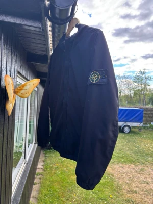 Svart bomberjacka från Stone Island - Snygg svart bomberjacka från Stone Island med klassisk patch på vänster ärm. Jackan har dragkedja framtill, ribbade muddar och en stilren design. Perfekt för dig som vill ha en ikonisk streetwear-look.