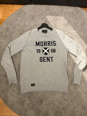 Ljusgrå stickad tröja Morris Gents - Ljusgrå stickad tröja från Morris Gents med svart text och tryck framtill. Tröjan har rund halsringning, ribbstickade muddar och nederkant. Perfekt för en stilren och avslappnad look.