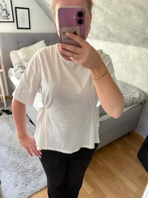 Vit topp med knyt i sidan - Snygg vit topp med knyt i sidan. T-shirtrmad och rund halsringning, perfekt för en clean och fräsch look. Tillverkad i mjuk bomull som känns skön mot huden.