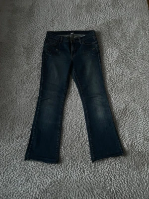 Vintage blå bootcut jeans med nitar - Snygga blå bootcut jeans från Oviesse med coola nitar och dekorativa detaljer framtill. Jeansen har klassisk femficksdesign och lätt utsvängda ben. Perfekta för dig som vill ha en unik touch på din denimstil. Köpta second hand i Paris. För kort för mig som är 168.