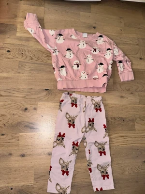 Rosa stretchbyxor med snögubbar och rådjur - Supergullig tvådelad pyjamas i ljusrosa med snögubbar på tröjan och rådjur med röda rosetter på byxorna. Mjukt material och bekväm passform, perfekt för mysiga vinterkvällar.