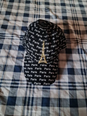 Svart keps med Paris-tryck och Eiffeltorn - Svart keps med vit text där det står 'Paris' över hela kepsen och ett stort guldfärgat Eiffeltorn broderat framtill. Klassisk böjd skärm och justerbar baksida. Perfekt accessoar för dig som gillar streetstyle och Paris-vibes.