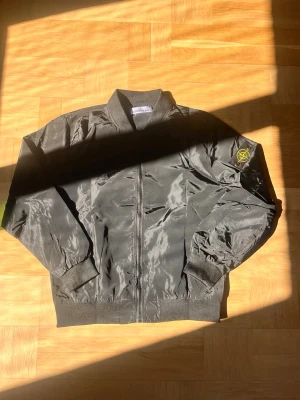Svart bomberjacka från Stone Island - Snygg svart bomberjacka från Stone Island med klassisk patch på vänster ärm. Jackan har ribbade muddar, dragkedja framtill och en stilren, minimalistisk design. Perfekt för dig som gillar streetwear och vill ha en ikonisk look.