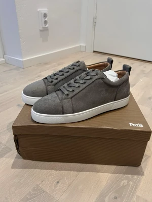 Gråa sneakers från Christian Louboutin - Stilrena grå/bige sneakers i mocka från Christian Louboutin med vita sulor och matchande grå skosnören. Klassisk låg modell med diskret design och bruna detaljer på insidan. Perfekta för dig som gillar exklusiva och minimalistiska skor.