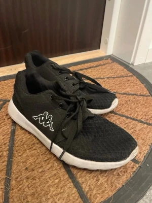 Svarta Kappa sneakers med vit sula - Svarta sneakers från Kappa med vit sula och klassisk logga på sidan. Ovandelen är i meshmaterial för bra andningsförmåga och skorna har svarta skosnören. Perfekta för en sportig och avslappnad stil.