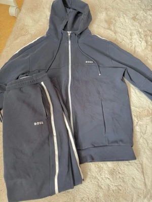 Mörkblå BOSS loungewear set - Snyggt mörkblått loungewear set från BOSS med vit dragkedja och vita ränder längs ärmar och byxor. Setet består av en hoodie med fickor och matchande byxor med resår i midjan. Perfekt för chill dagar hemma. Materialet känns mjukt och bekvämt.