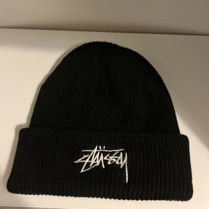 Svart Stüssy mössa - Svart ribbstickad mössa från Stüssy med vit broderad logga framtill. Klassisk uppvikt kant och mjukt material som håller dig varm. Perfekt för dig som gillar streetwear och vill ha en clean look.