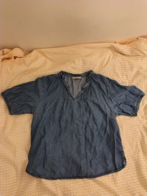 Blå jeansblus/topp - Snygg blå jeansblus med v-ringad hals och dekorativa sömmar vid halsringningen. Blusen har korta ärmar och är tillverkad i ett mjukt denimliknande material. Perfekt för en avslappnad och vintage eller trendig, stilren look. Endast använd ett fåtal gånger. 