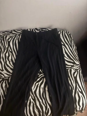 Svarta kostymbyxor med kritstrecksrand - Snygga svarta kostymbyxor med smala vita kritstrecksränder. Klassisk rak passform och hällor för bälte. Perfekta för en stilren look. Materialet ser ut att vara en blandning av polyester och viskos.