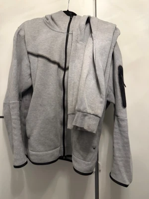 Nike tech tracksuit - Säljer en Nike tracksuit  med hel dragkedja och svarta detaljer. Hoodien har en huva, fickor framtill och en extra ficka med dragkedja på ena ärmen. Tillverkad i mjukt sweatshirtmaterial, perfekt för chill dagar. Och byxorna perfekt för vardagen och träning.