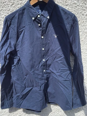 Mörkblå skjorta från Gant - Klassisk mörkblå skjorta från Gant med vit knappstängning och broderad logga på bröstet. Skjortan har lång ärm, krage och är tillverkad i bomull. Perfekt för en stilren och avslappnad look.