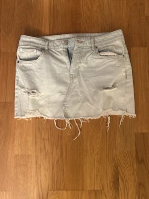 Ljus lågmidjad jeanskjol med slitningar - Säljer en ljusblå jeanskjol med slitna detaljer och fransig nederkant. Kjolen har klassiska fem fickor och råa kanter för en trendig look. Perfekt för dig som gillar en avslappnad och cool stil. Skitsnygg har dock en fläck men är inget som märks på!! Pris kan diskuteras storleken passar ungefär S-M