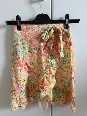 Blommig omlottkjol med volang - Söt kort omlottkjol med volangkant och knytband i midjan. Kjolen har ett färgglatt blommigt mönster i gult, grönt, rosa och rött. Kjolen känns lätt och somrig, perfekt för varma dagar.
