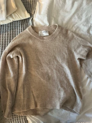 Beige stickad tröja från Selected Femme - Mysig beige stickad tröja från Selected Femme med rund halsringning och ribbade muddar. Tröjan har en avslappnad passform och är tillverkad i ett mjukt material som passar perfekt för kyliga dagar.