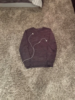 Ralph Lauren Tröja Grå - Mörkgrå stickad tröja från Polo Ralph Lauren med rund halsringning och diskret logga broderad på bröstet. Tröjan har en klassisk passform och är tillverkad i ett mjukt, melerat material. Perfekt för lager-på-lager-stil.