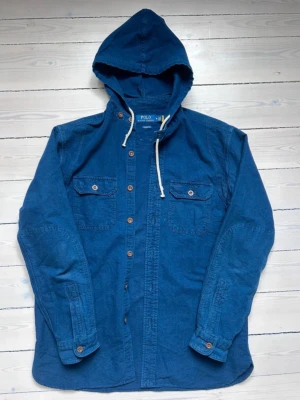 Blå overshirt med huva från Polo Ralph Lauren - Snygg blå overshirt från Polo Ralph Lauren med huva och vita dragsnören. Skjortan har två bröstfickor med lock och knappar samt knäppning hela vägen fram. Tillverkad i ett mjukt bomullstyg, perfekt för lager-på-lager-stil.