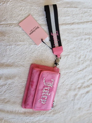 Juicy Couture rosa plånbok med strass - Säljer en fluffig rosa plånbok från Juicy Couture med glittrande strassdetaljer på framsidan. Plånboken har dragkedja, ficka framtill och en bred svart och vit handledsrem med rosa Juicy-logga. Perfekt accessoar för dig som gillar statement pieces.