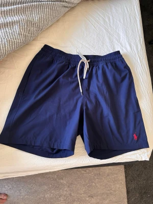 Marinblå shorts från Polo Ralph Lauren - Snygga marinblå shorts från Polo Ralph Lauren med elastisk midja och vit dragsko. Klassisk röd logga broderad framtill och praktisk bakficka med knapp. Perfekta för sommaren och chill dagar. Materialet känns lätt och bekvämt.