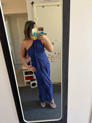 Blå oneshoulder långklänning - Snygg blå långklänning med oneshoulder-design och draperad knut framtill. Klänningen har en elegant och flytande passform som ger en stilren siluett. Perfekt för dig som vill sticka ut med en modern och trendig look.Helt ny Storlek 38/40 märkt som L