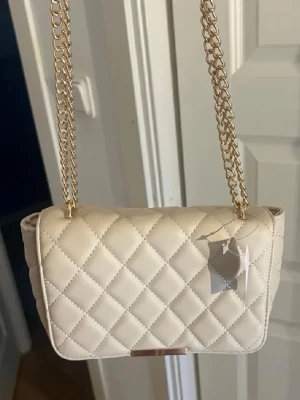 Beige quiltad axelväska med guldkedja - Snygg beige axelväska med quiltat mönster och guldfärgad kedjerem. Väskan har en klassisk rektangulär form och stängs med lock. Perfekt för dig som gillar stilrena accessoarer med lyxig känsla. Materialet ser ut att vara konstläder.