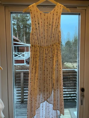 Vit barnklänning med blommigt mönster - Vit långklänning med små blommor i olika färger. Klänningen är ärmlös och har en lätt, luftig känsla med resår i midjan och asymmetrisk nederkant. Perfekt för varma dagar och somriga tillfällen.