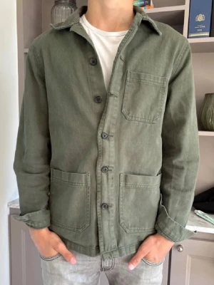Grön Overshirt  - Stilren grön overshirt i kraftigt bomullstyg med tre stora fickor framtill och knäppning med svarta knappar. Klassisk krage och raka ärmar, perfekt att slänga över en t-shirt för en avslappnad look.