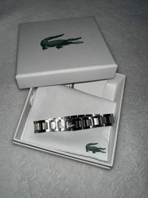 Silverfärgat armband från Lacoste - Säljer ett Lacoste armband i storlek M som endast är användt ett fåtal gånger, den kommer i orginalförpackningen och är i perfekt skick!