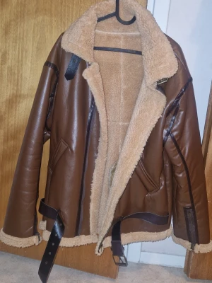 Teddyjacka/Bomberjacka med bälte och fuskpäls - Snygg brun teddyjacka med mjukt beige fuskpälsfoder på insidan och en krage. Jackan har svarta detaljer, bälte i midjan, samt vid halsen om man inte vill ha kragen nervikt. Dragkedja finns vid ärmslut samt två fickor framtill. Perfekt för kalla dagar och ger en cool vintage vibe. Sparsamt använd, finns ingen lapp men passar en M/liten L.
