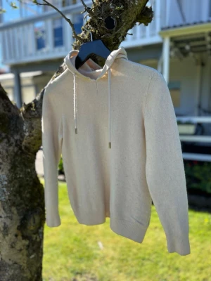 Beige merinoull hoodie med luva - Snygg beige stickad hoodie med huva och dragsko. Tröjan har långa ärmar och ribbade muddar vid ärmslut och nederkant. Perfekt för en chill och avslappnad stil. Materialet känns mjukt och skönt mot huden.
